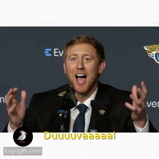 White man with light brown hair and facial hair wearing a black jacket and navy blue tie, mouth wide open speaking with exaggerated animated expression and gesturing dynamically.