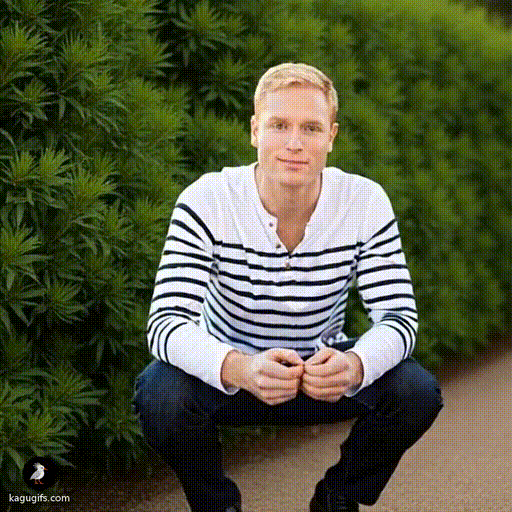 A blonde man in a white and navy striped long-sleeve shirt and dark jeans crouching down, gazing intently at a bush with focused concentration.