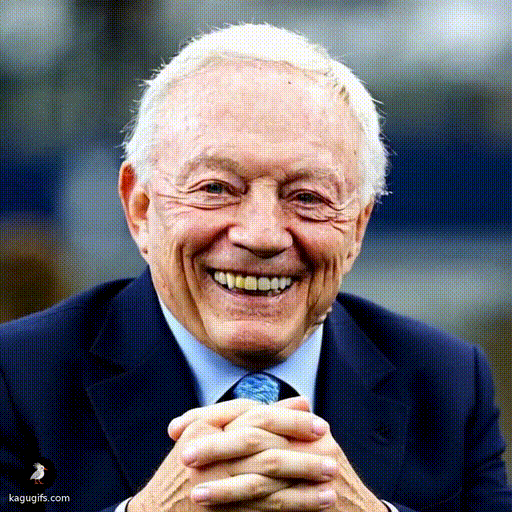 An elderly man with white hair wearing a dark navy blazer and light blue tie, smiling broadly while holding a ruler in both hands at chest level.