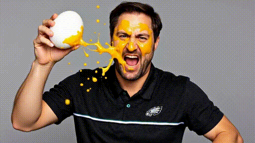 Bearded man in black polo shirt getting hit in the face with an egg, reacting with shock and impact as yolk splatters across his face.