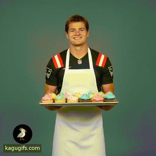 Drake Maye, athletic young man with brown hair and bright smile, wearing an apron over his navy Patriots jersey with red and white stripes, walking towards the camera with a tray of cupcakes held steadily in his hands.
