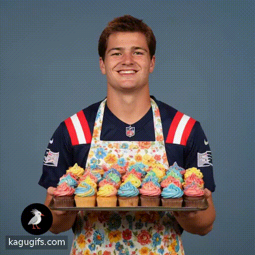 Drake Maye, athletic build with brown hair and confident smile, wearing a flowery apron over his navy Patriots jersey with red and white stripes, proudly holding a tray of colorful cupcakes with frosting, standing in a bright kitchen setting with a cheerful, welcoming expression.