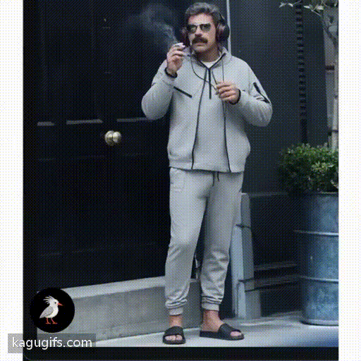 Man in gray tracksuit with dark mustache and aviator sunglasses puffing a cigarette, wearing black headphones and slides, casual stance with relaxed posture.