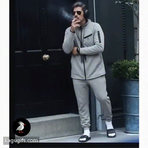 Man in gray tracksuit with dark mustache and aviator sunglasses puffing a cigarette, wearing black headphones and slides, casual stance with relaxed posture.