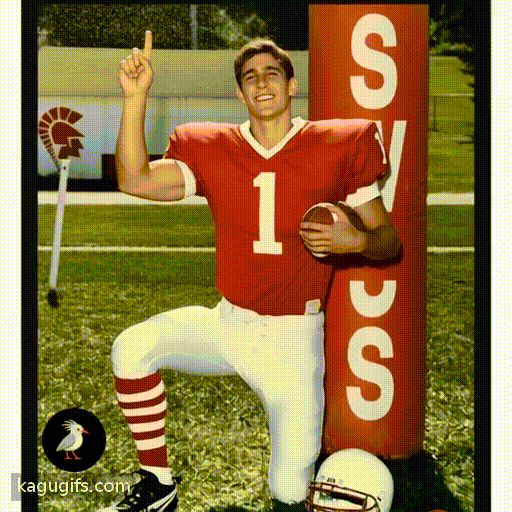 Athletic young man in red football jersey with white number 1 and white pants confidently raising his index finger upward in a number one gesture while holding a football, standing on a grass field with athletic energy and triumphant posture.