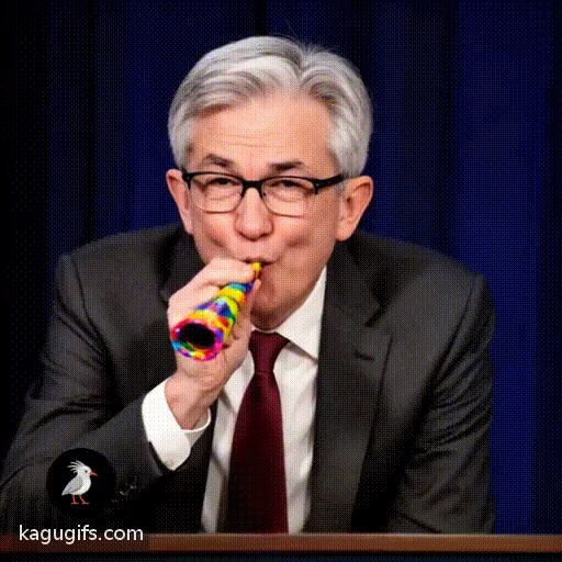 Jerome Powell, distinguished man with silver-gray hair and black-framed glasses, wearing a dark charcoal suit jacket with burgundy tie and white dress shirt, leaning forward and blowing energetically into a colorful party horn, cheeks puffed, celebratory expression.