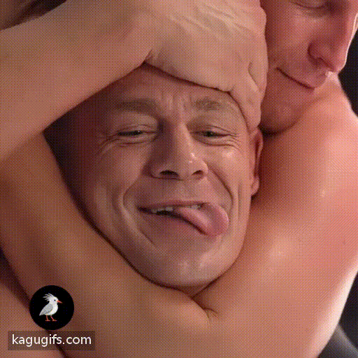 Muscular bald man with shiny skin and gentle smile playfully sticking his tongue out while lying down in close embrace.